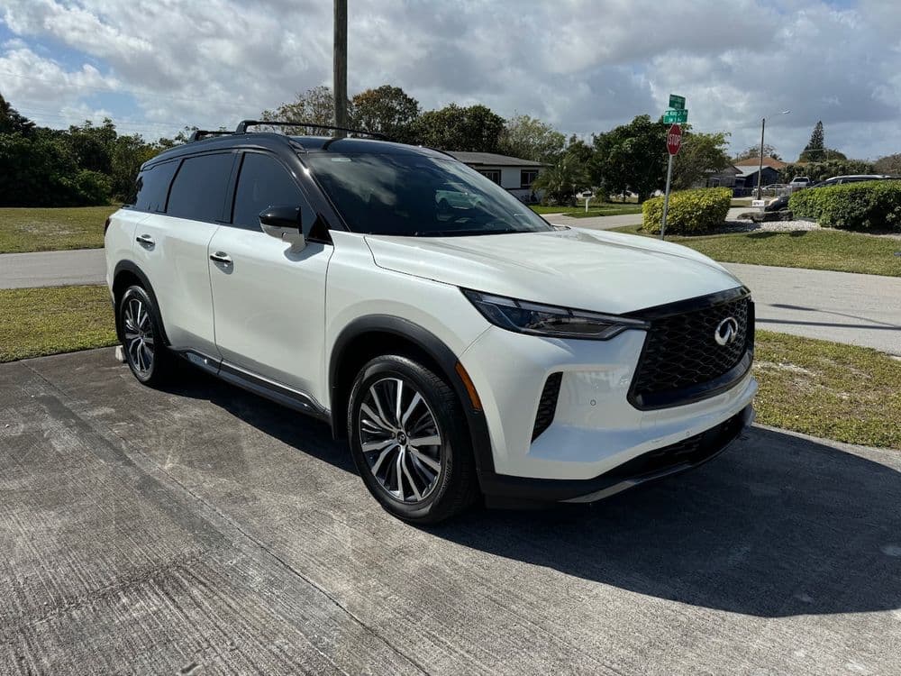 White Infiniti QX60 parked on a sunny street with green landscaping in the background.