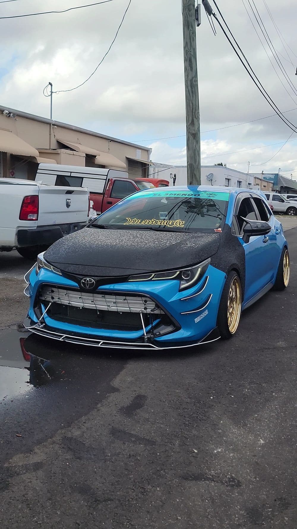 Modified blue Toyota car with carbon fiber hood and gold wheels parked on a street.