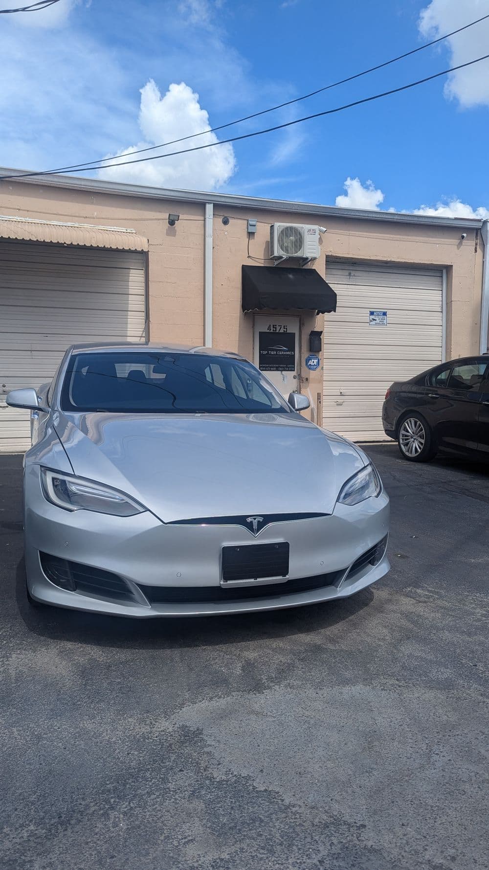 Silver Tesla Model S parked in front of a garage with blue sky and clouds overhead.