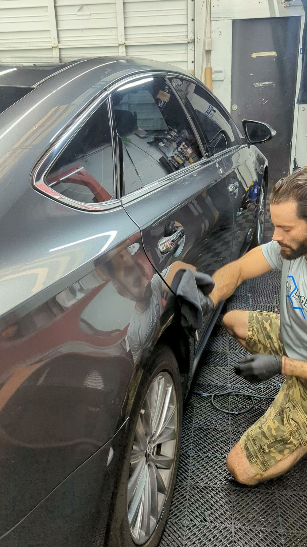 Car detailing process with technician cleaning a gray sedan's rear panel in a garage workshop.