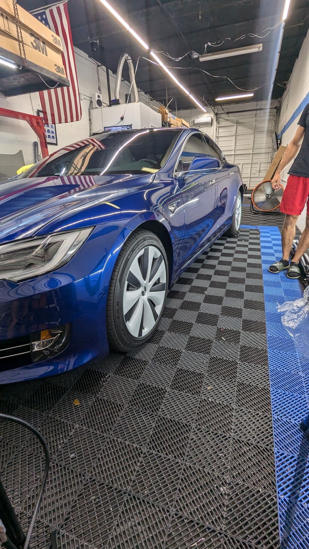 Blue electric car on checkered floor in a garage, with technician working nearby.