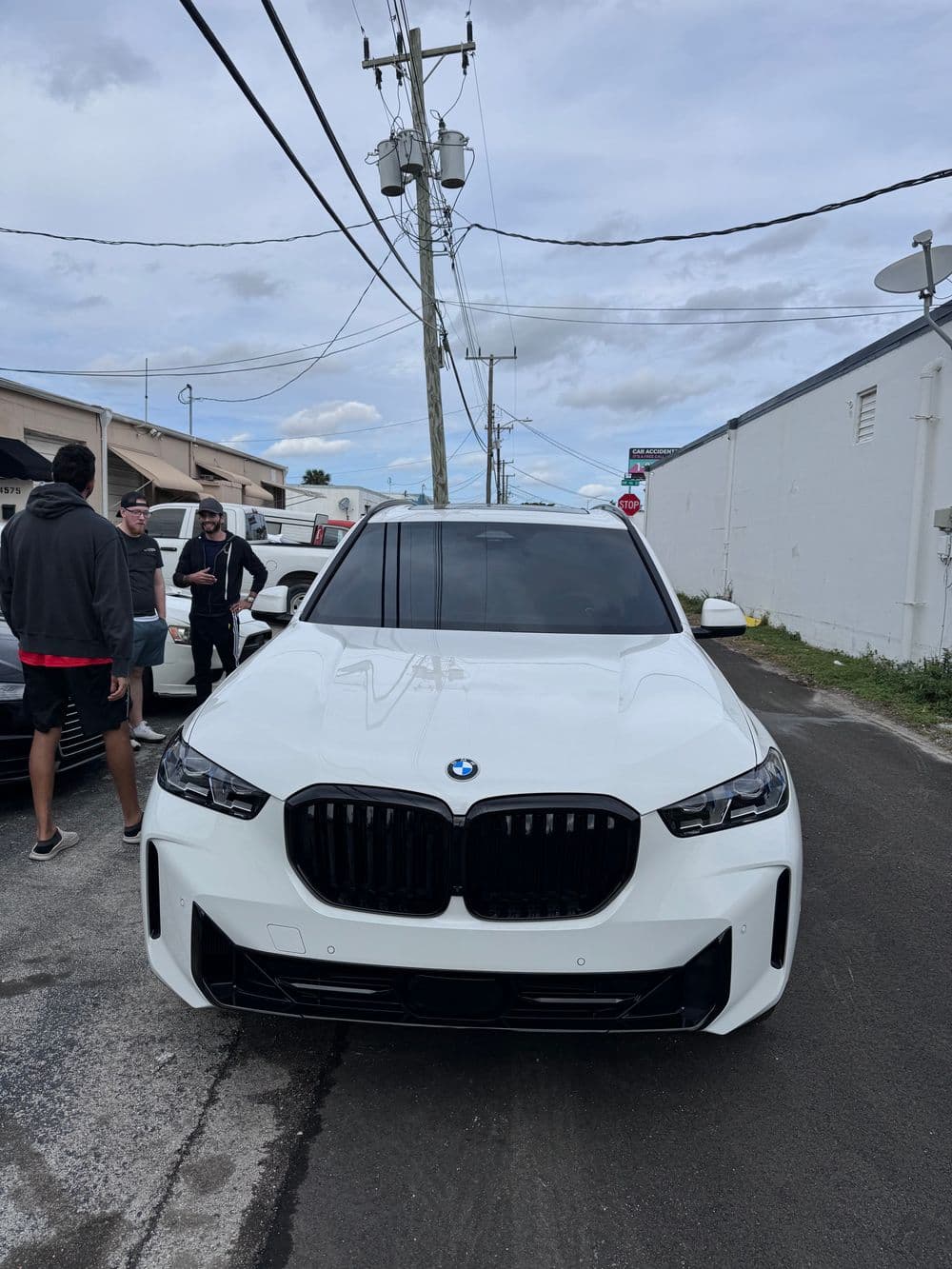 Front view of a white BMW X7 in an urban setting with people and power lines in background.