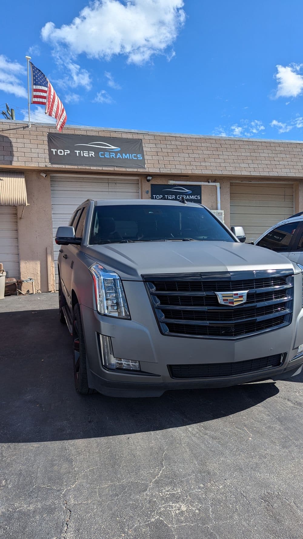 Matte gray Cadillac SUV parked outside Top Tier Ceramics with American flag in background.