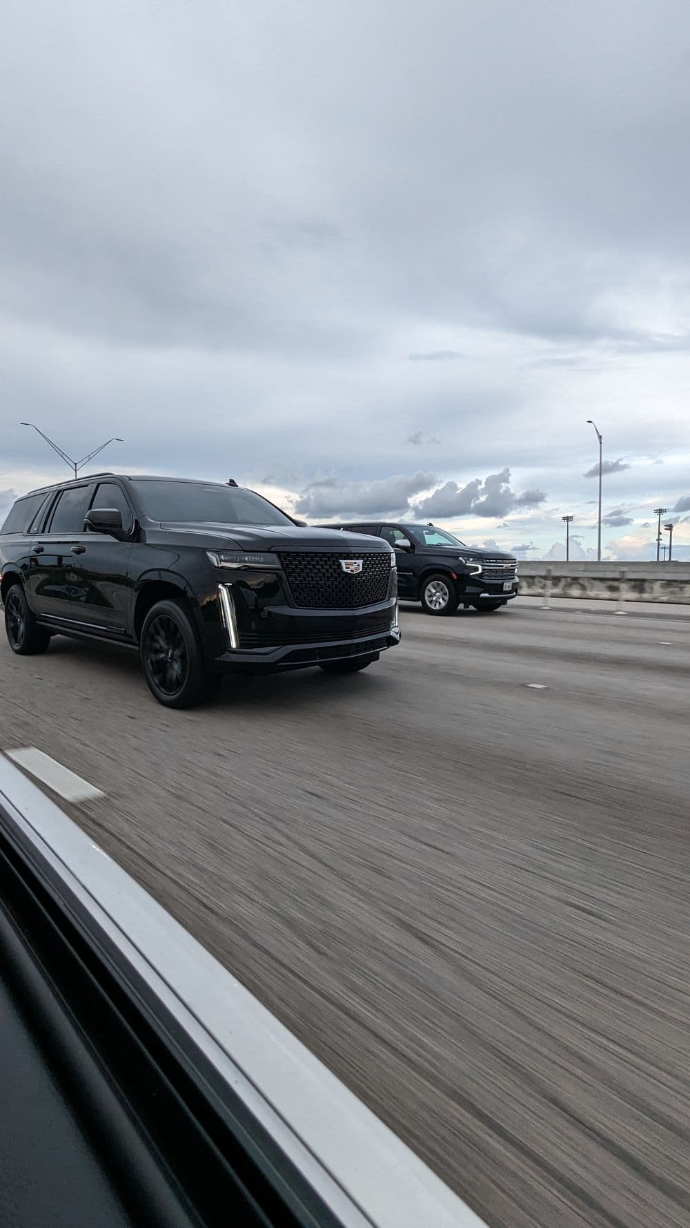 Two black SUVs on a highway under a cloudy sky, showcasing modern vehicle design.