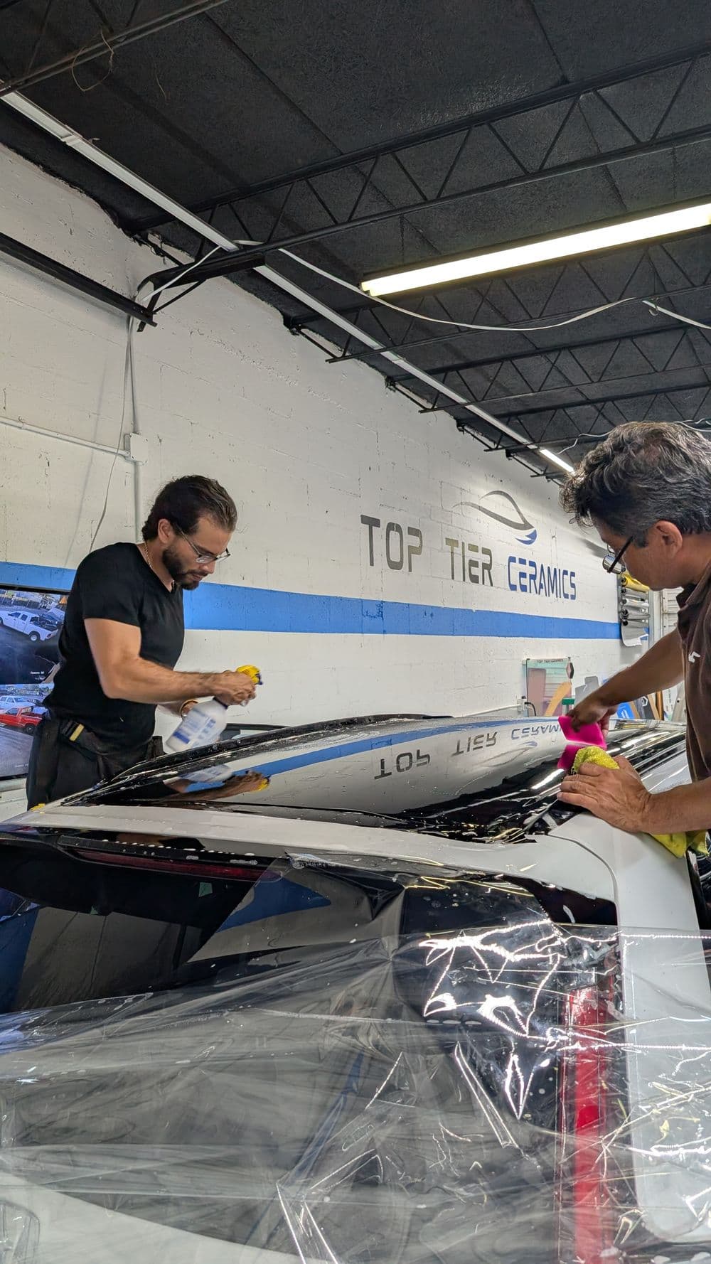 Two technicians applying ceramic coating on a car roof in a detailing shop.